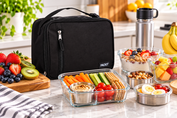 Black PackIt freezable lunch bag beside containers of fresh fruit, vegetable sticks, yoghurt and snacks on a kitchen counter.