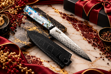 Premium Damascus kitchen knife with a colourful patterned handle and black leather sheath on burgundy and white marble with luxurious gold accents.