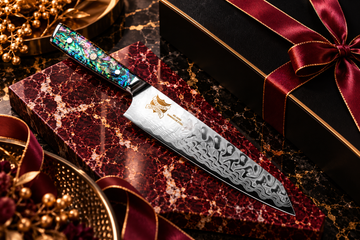 Iridescent-handled chef’s knife on burgundy marble with a black gift box and gold accents.