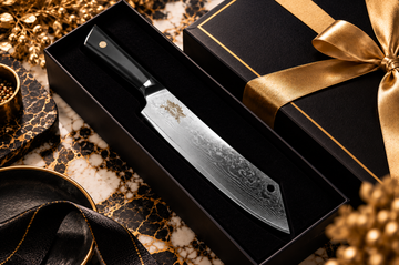 Patterned chef’s knife in a black presentation box with gold ribbon on a black and gold marble surface.