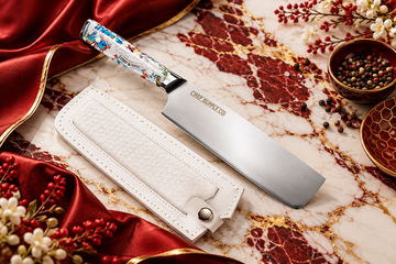 Premium Chef Supply Co. kitchen cleaver with a colourful handle and white leather sheath on white marble with burgundy red and gold accents.